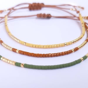 Pode incluir: Três pulseiras sobrepostas com contas douradas, verdes e marrons. As pulseiras são amarradas com cordões marrons.