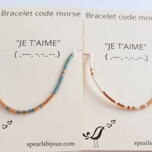 Op de afbeelding: Twee armbanden met de boodschap "JE T'AIME" in morsecode. De armbanden zijn gemaakt van kleine kralen in teal en wit met gouden accenten. De armbanden worden getoond op kaarten met de boodschap en de morsecode vertaling.