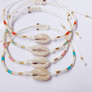 Puede incluir: Cuatro pulseras de cordón blanco con abalorios de conchas marinas. Cada pulsera presenta cuentas de colores con detalles en rojo, rosa, azul y dorado. Las pulseras son ajustables y están anudadas.