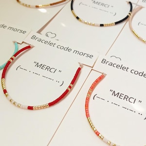 Peut inclure: Une collection de bracelets fins composés de perles colorées et d'accents dorés, montés sur des cartes blanches portant l'inscription Bracelet code morse, le mot MERCI et la séquence en code Morse correspondante imprimés en noir.