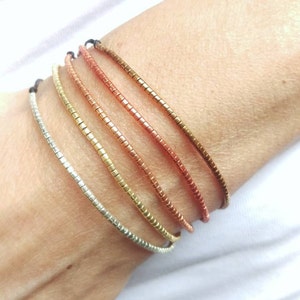 May include: Five stacked bracelets made of small metal squares. The bracelets are in shades of silver, gold, and copper.