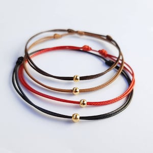 Könnte beinhalten: Drei Freundschaftsarmbänder mit goldenen Perlen. Die Armbänder sind rot, braun und schwarz.