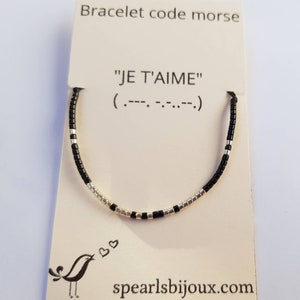 Può includere: Un braccialetto di perline nere e argentate con la frase "JE T'AIME" e la sua traduzione in codice Morse su un cartoncino bianco. Il cartoncino presenta anche una piccola illustrazione di un uccello e il sito web "spearlsbijoux.com".