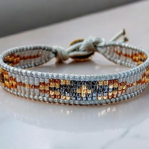 Peut inclure: Bracelet fait à la main avec un motif tissé. Le bracelet présente un motif de perles argentées, dorées et noires. Le bracelet est réglable et présente une palette de couleurs neutres.
