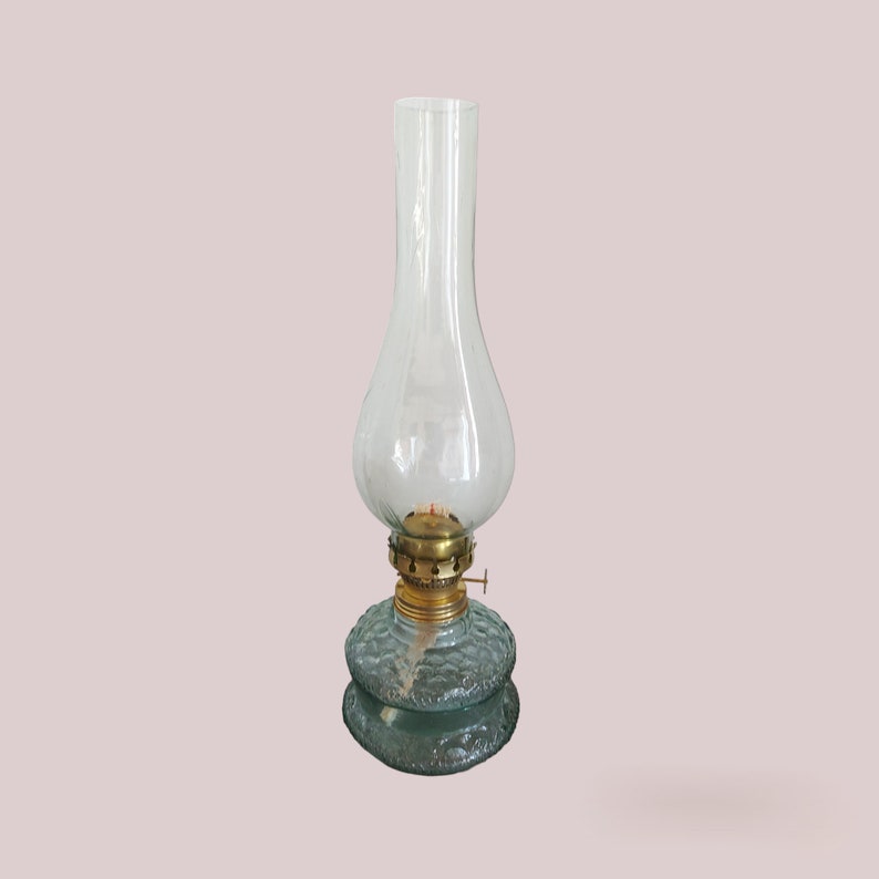 May include: A clear glass oil lamp with a brass wick adjuster and a textured glass base. The lamp is on a light pink background.