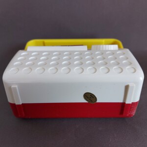 May include: A white and red vintage toy radio with a yellow top. The radio has a round knob on the top and a sticker that says "Made in Hong Kong".