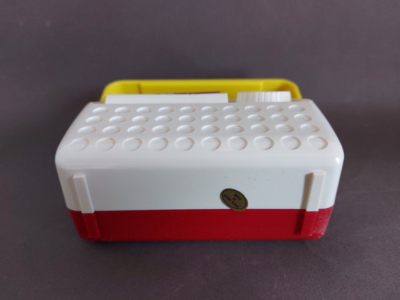 May include: A white and red vintage toy radio with a yellow top. The radio has a round knob on the top and a sticker that says "Made in Hong Kong".