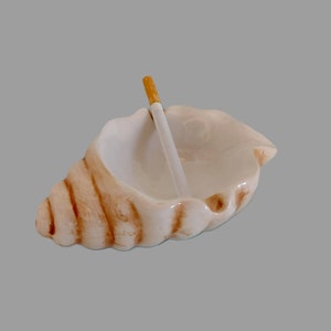 Op de afbeelding: Een witte keramische schelpvormige asbak met een bruin swirl-ontwerp. Een sigaret rust in de asbak.
