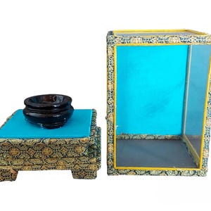 Puede incluir: Un cuenco negro descansa sobre un cuadrado turquesa encima de un soporte decorativo de varios niveles. El soporte y una caja a juego con los lados abiertos presentan una tela estampada dorada y verde. La caja tiene un interior turquesa y ribetes amarillos.