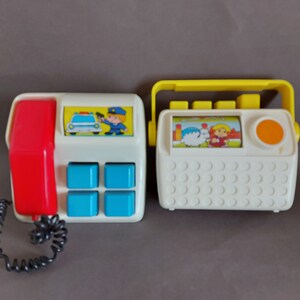 May include: Two vintage toy phones with a white base and colorful accents. One phone has a red receiver and blue buttons, and the other has a yellow handle and an orange button. Both phones feature cartoon images.