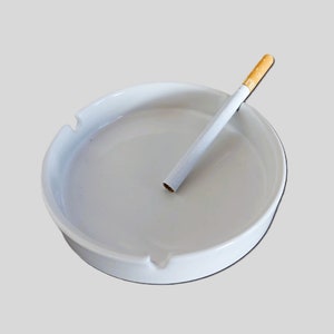 Pode incluir: Um cinzeiro redondo de cerâmica branca com um cigarro. O cinzeiro tem uma borda ligeiramente elevada e um entalhe para apoiar os cigarros. O cigarro é branco com um filtro castanho.
