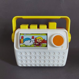 May include: A white and yellow toy radio with a red and white picture of a girl and a sheep. The radio has a yellow handle and a round orange button.