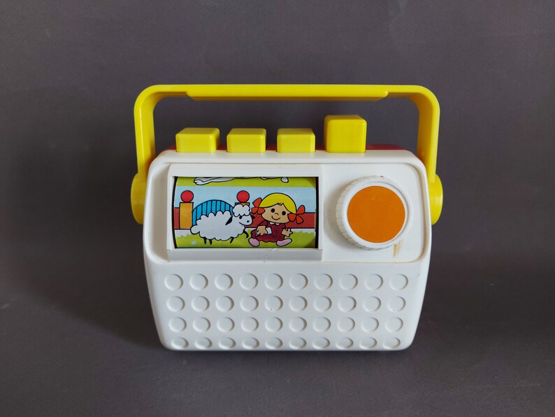 May include: A white and yellow toy radio with a red and white picture of a girl and a sheep. The radio has a yellow handle and a round orange button.