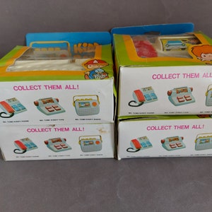 May include: Four boxes of vintage toy phones, each with a different design. The boxes are white with colorful illustrations of the phones and the text "COLLECT THEM ALL!" The boxes are stacked on top of each other.