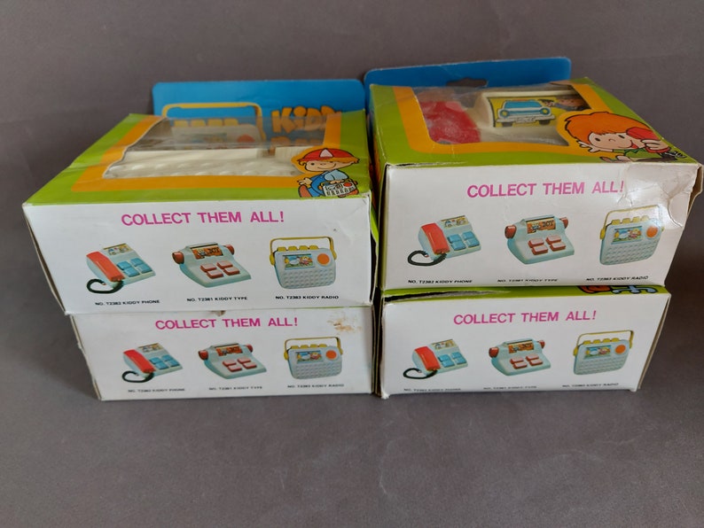 May include: Four boxes of vintage toy phones, each with a different design. The boxes are white with colorful illustrations of the phones and the text "COLLECT THEM ALL!" The boxes are stacked on top of each other.