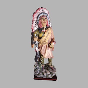 May include: A figurine of a Native American man wearing a colourful headdress and holding a pipe. He is standing on a rocky base with green foliage.