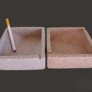 Puede incluir: Dos ceniceros cuadrados de color marrón claro y moteados, con un cigarrillo apoyado en uno de ellos.