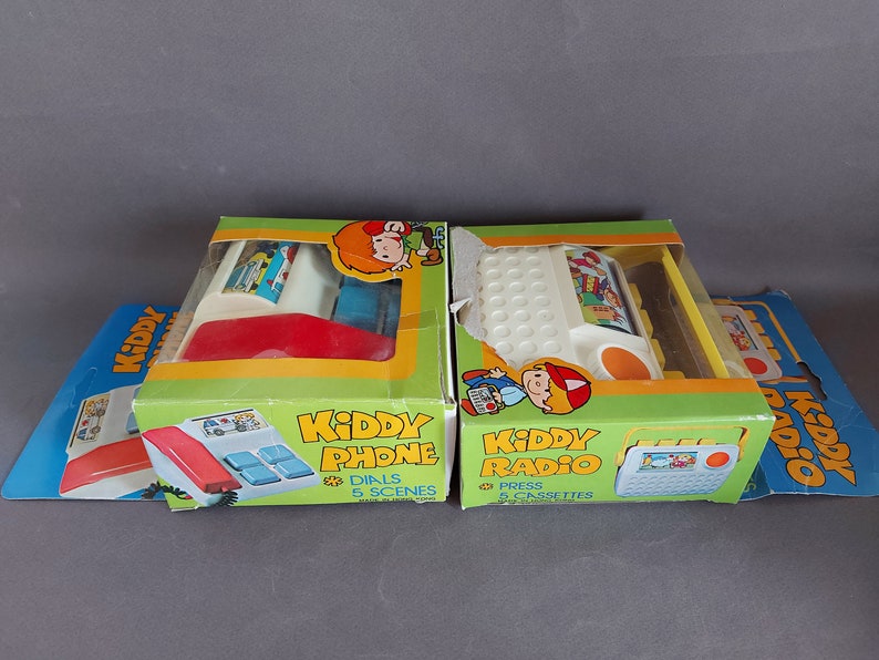 May include: Two vintage toy boxes with Kiddy Phone and Kiddy Radio on the front. The phone is red and white with a dial and the radio is white and yellow with a cassette player. Both boxes have cartoon characters on them.