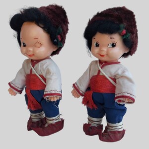 May include: Two vintage dolls wearing traditional clothing with red sashes and brown fur hats. The dolls have black hair and painted faces.