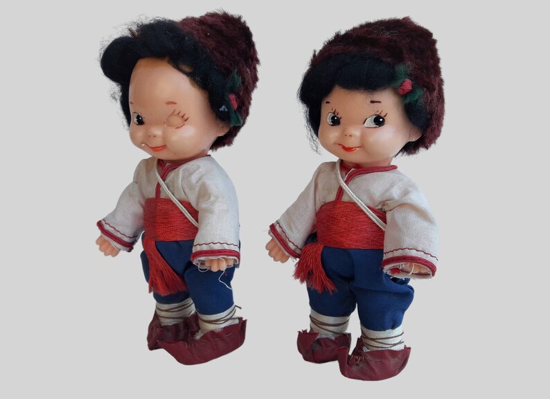 May include: Two vintage dolls wearing traditional clothing with red sashes and brown fur hats. The dolls have black hair and painted faces.