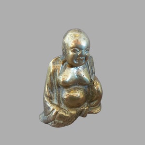 May include: A small, antique-looking brass figurine of a seated, smiling Buddha. The figure has a rounded belly and is wearing draped robes. The metal has a slightly tarnished appearance.