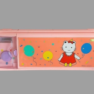 Peut inclure: Une trousse à crayons rose Hello Kitty avec une fenêtre en plastique transparent et un motif Hello Kitty avec des ballons et des confettis. La trousse a un bouton violet et le texte "MKO-0101" en bas.