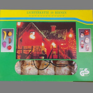 Op de afbeelding: Een doos voor een kerstverlichtingssnoer met 10 lampjes, met kerstmanfiguren en andere kerstfiguren. De doos heeft een groene achtergrond met een gele rand en de tekst "Lichterkette 10 Birnen" bovenaan. De doos bevat ook een afbeelding van een huis dat versierd is met kerstverlichting.