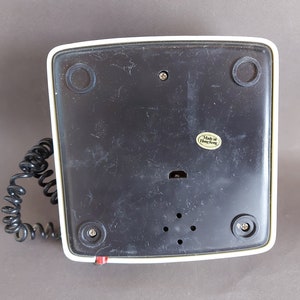 May include: The bottom of a white and black rotary phone with a coiled cord. The phone has a black plastic base with a label that reads "Made in Hong Kong".