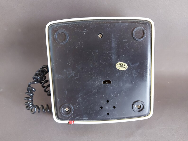 May include: The bottom of a white and black rotary phone with a coiled cord. The phone has a black plastic base with a label that reads "Made in Hong Kong".