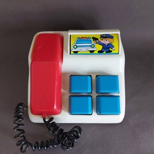 May include: A white toy telephone with a red receiver and a blue and yellow police car image. The telephone has four blue buttons and the text "POLICE" on the car image.