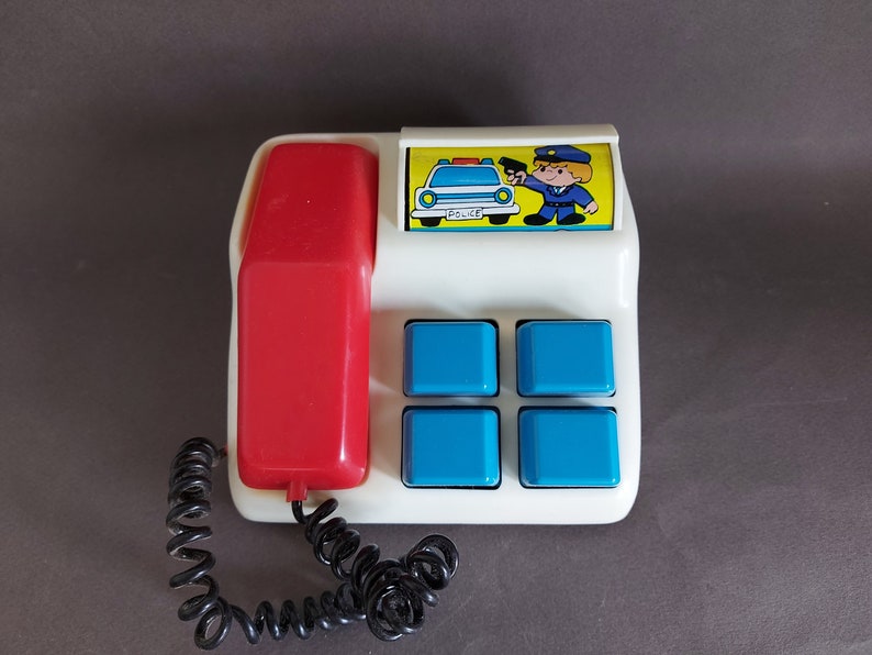 May include: A white toy telephone with a red receiver and a blue and yellow police car image. The telephone has four blue buttons and the text "POLICE" on the car image.