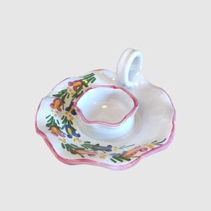 May include: White ceramic candle holder with a pink rim and floral design. The holder has a small, round well for a candle and a handle on the side.