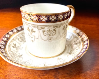 Puede incluir: Una taza de té y un platillo blancos con un borde dorado y azul. La taza y el platillo tienen un delicado patrón floral en oro.