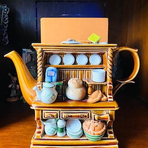 May include: A novelty ceramic teapot in the form of a kitchen dresser, with shelves displaying miniature plates, cups, and food. The teapot is predominantly brown and yellow, with green and white details. Includes a small teapot and a "Caster Sugar" container.