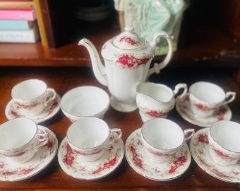 May include: A vintage tea set with a white teapot, creamer, and six teacups and saucers. The set features a floral pattern with red and gold accents.