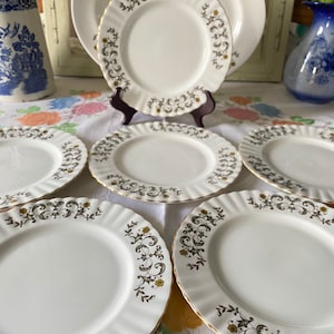 May include: Set of white ceramic dinner plates with a gold rim and a green and gold floral pattern. The plates are arranged in a circle on a white tablecloth.
