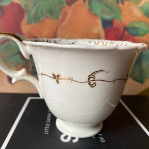 May include: A white teacup with a gold rim and a gold floral design. The teacup is sitting on a black surface.