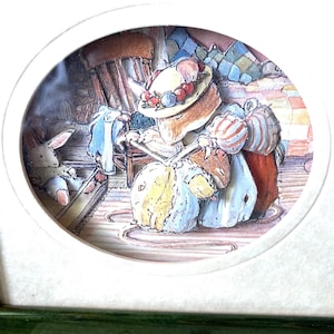 Puede incluir: Una ilustración redonda enmarcada de un ratón con un sombrero y un vestido, sentado en una silla y tejiendo. Un conejo blanco está de pie en un escalón de madera en el fondo. El marco es verde y tiene el texto "BRAMBLEHEDGE" en la parte inferior.