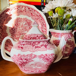 Puede incluir: Una tetera de cerámica blanca y roja con un diseño de paisaje. También se ven una fuente grande y una jarra a juego. Flores blancas en un jarrón en el fondo. El juego es probablemente vintage.