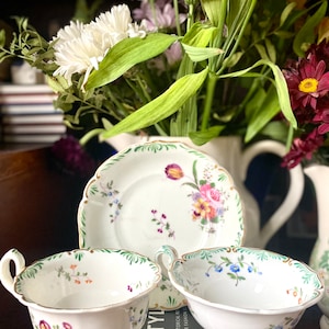 Puede incluir: Dos tazas de té blancas con ribetes verdes y dorados y patrones florales, y un platillo a juego con un patrón floral más grande. Las tazas y el platillo están sobre un libro negro con la palabra "STYLE" visible.