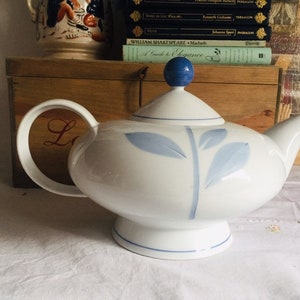 May include: A white teapot with a blue floral design and a blue knob on the lid. The teapot is sitting on a white tablecloth.