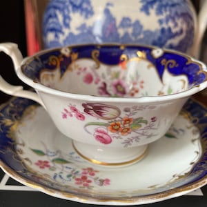 May include: A white teacup and saucer set with a floral design and gold trim. The rim and outer edge of the saucer are navy blue with gold accents. The teacup has a curved handle.