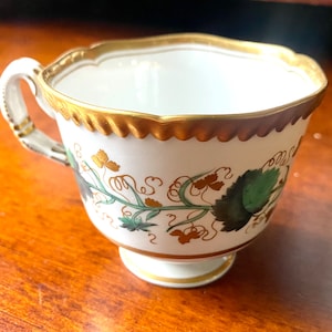 Puede incluir: Una taza de té blanca con un borde dorado y un diseño floral. La taza tiene un asa delicada y un pie pequeño.