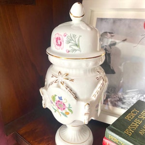 Könnte beinhalten: Weiße Keramikurne mit goldener Verzierung und Blumendesigns. Die Urne hat einen Deckel und steht auf einem Sockel. Die Urne steht auf einem Regal mit einem Stapel Bücher.