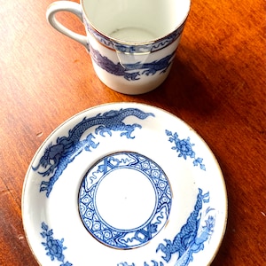 May include: A white porcelain teacup and saucer set with a blue and gold dragon design. The cup and saucer have a gold rim.
