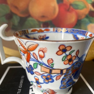 May include: A white porcelain teacup with a floral pattern in shades of blue, red, orange, and green. The cup has a unique handle and a gold rim.