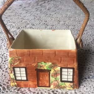May include: A brown ceramic cottage-shaped sugar bowl with a woven wicker handle. The bowl has a white interior and is decorated with a brick pattern, green vines, and two windows.