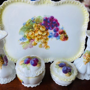 Könnte beinhalten: Ein weißes Porzellantablett mit einem Blumendesign mit Trauben und Erdbeeren. Das Tablett ist von vier weißen Porzellanstücken umgeben, zwei Kerzenhaltern und zwei kleinen runden Behältern mit Deckeln. Alle Teile sind mit Blumendesigns verziert.