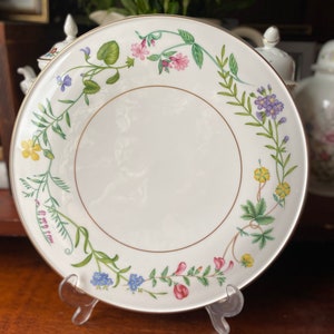 May include: A white porcelain plate with a gold rim and a floral design featuring pink, purple, yellow, and blue flowers. The plate is on a clear plastic stand.