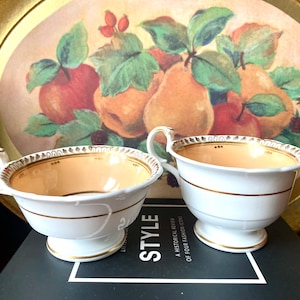 May include: Two white teacups with gold trim and a light orange interior. The cups are sitting on a black surface with a book that has the word "Style" printed on it. The cups are in front of a gold tray with a painted design of fruit and leaves.